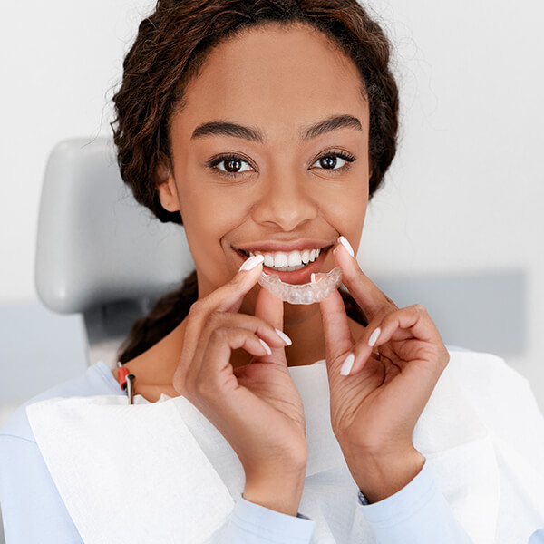 Young female sitting in a dentist’s chair and smiling is holding clear aligners.